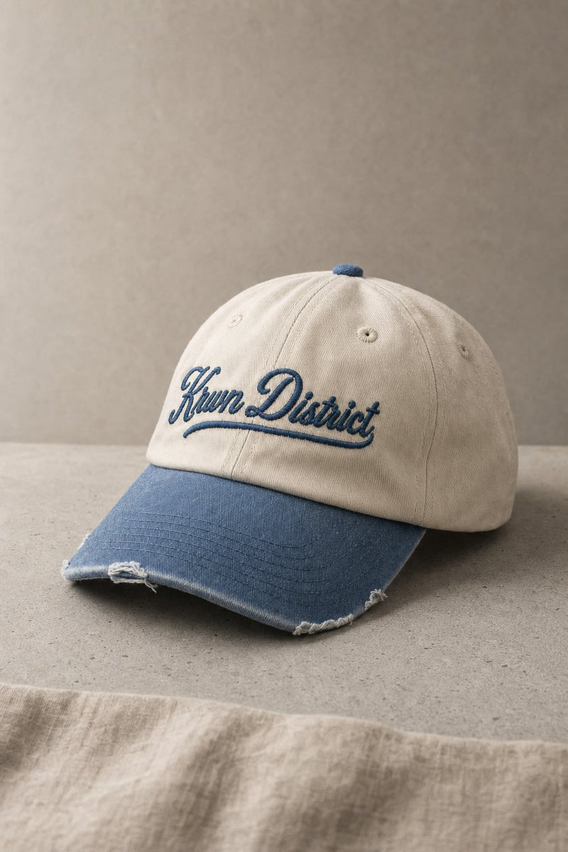 KRWN DISTRICT The Culture Two-Tone vintage dad cap — cream crown, washed indigo visor with frayed edge, three-quarter still life on concrete (AI-generated)