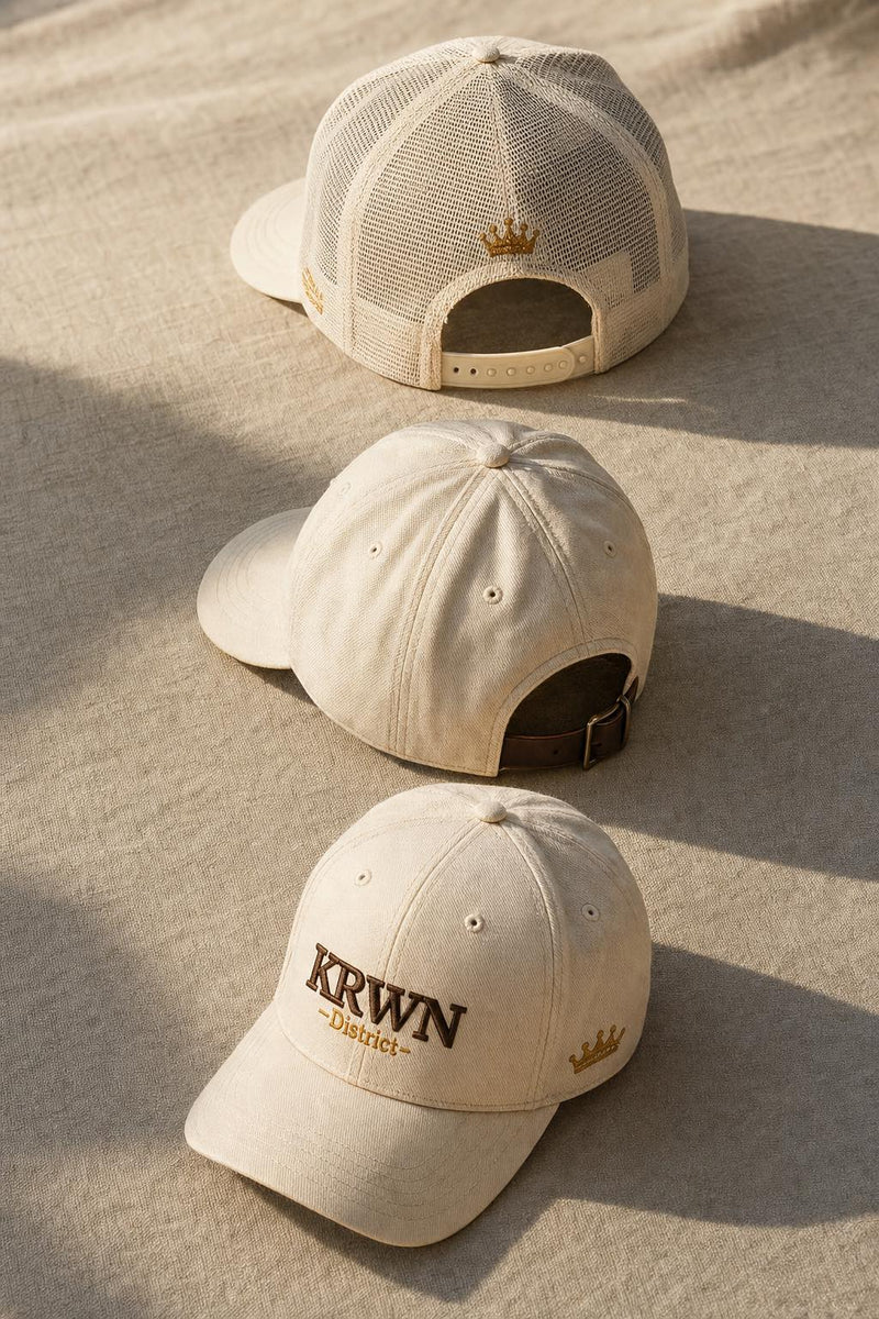 Three KRWN District premium caps arranged in editorial flat-lay — Trucker snapback, Baseball adjustable strap, and fitted closure styles compared side by side on warm linen surface.