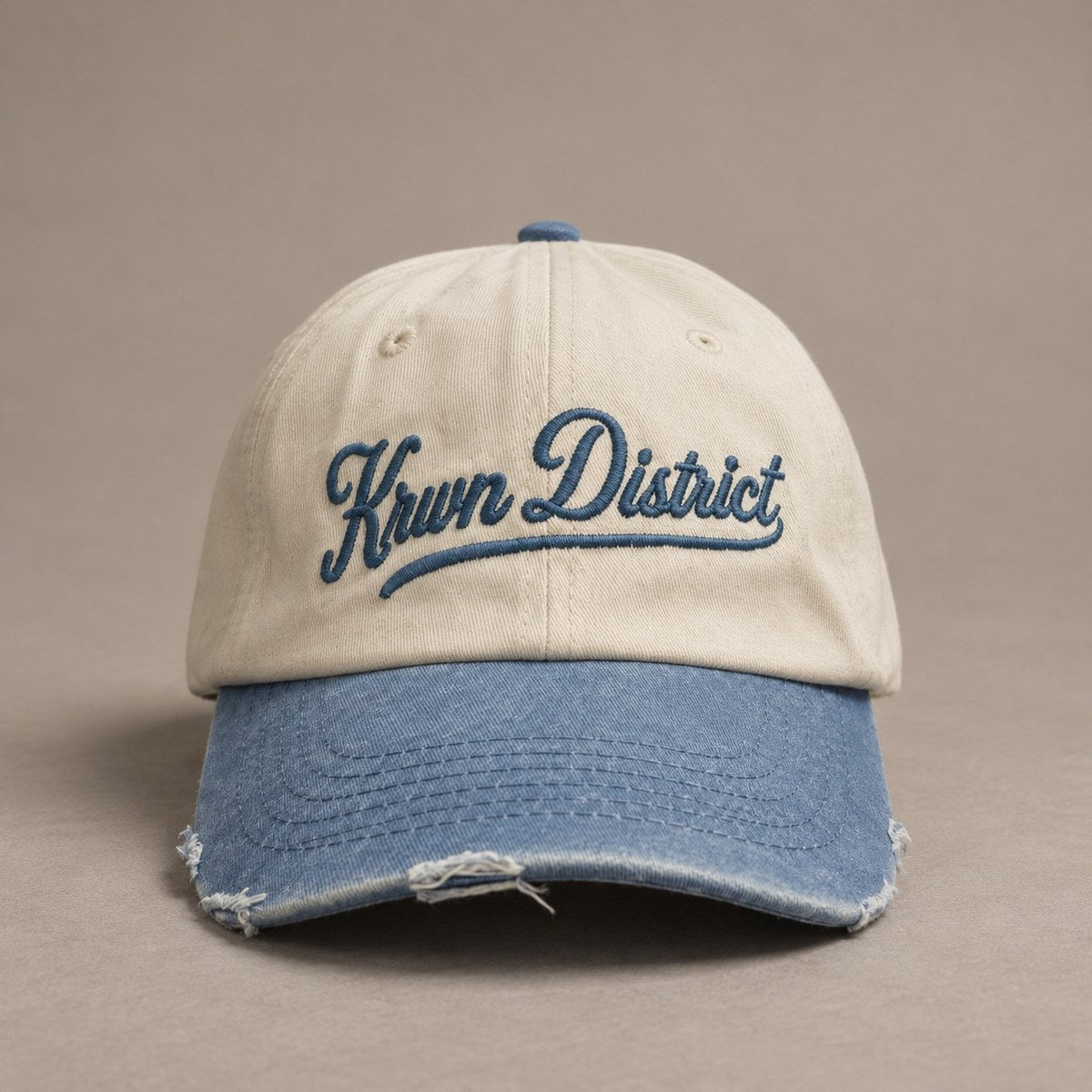 KRWN DISTRICT The Culture Two-Tone — vintage 6-panel dad cap, cream crown with washed indigo visor and blue Krwn District script embroidery, front view (AI-generated)