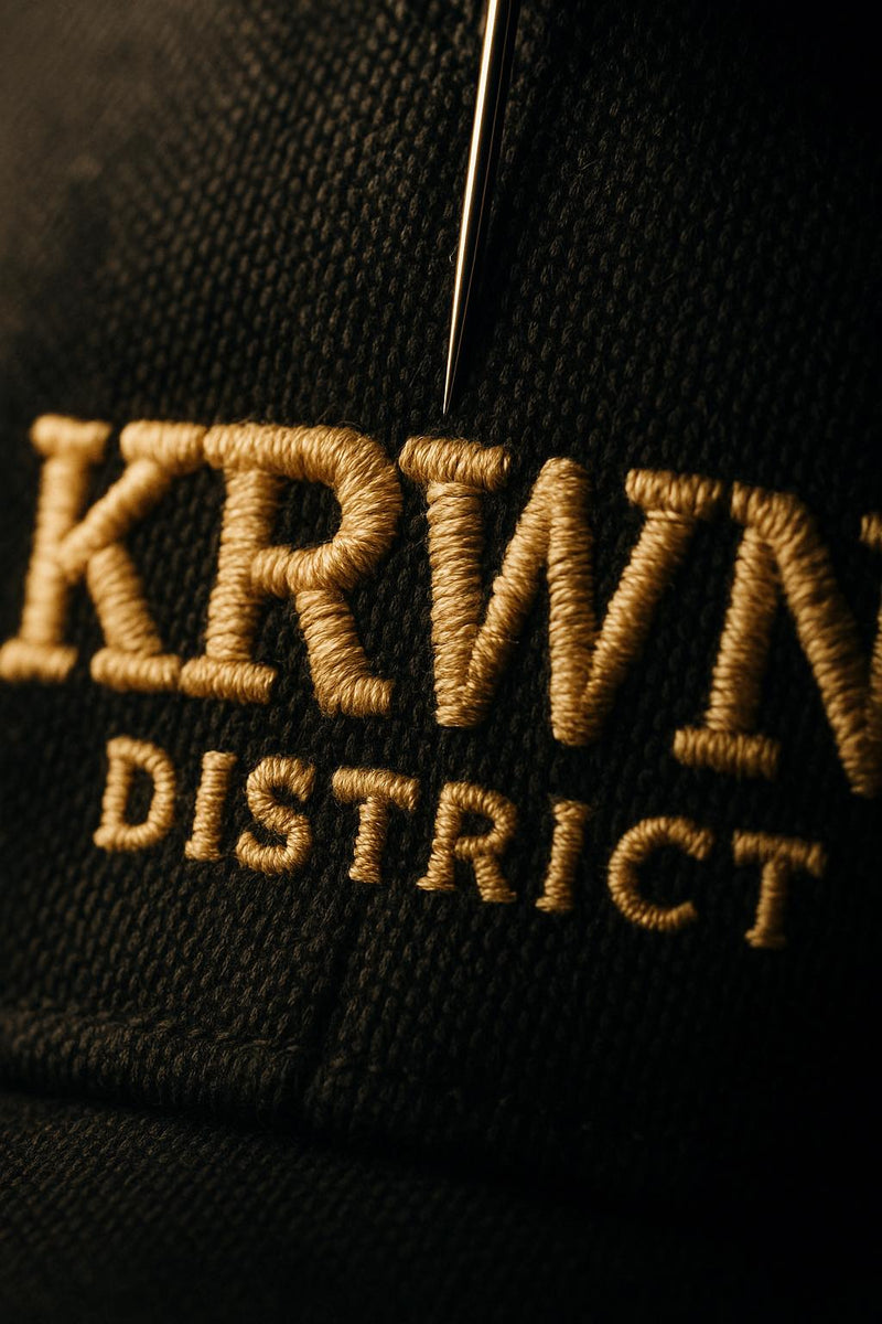 Macro close-up of embroidered KRWN District logo — golden cream thread sewn into heavy dark cotton canvas, needle visible mid-stitch under dramatic side-light.